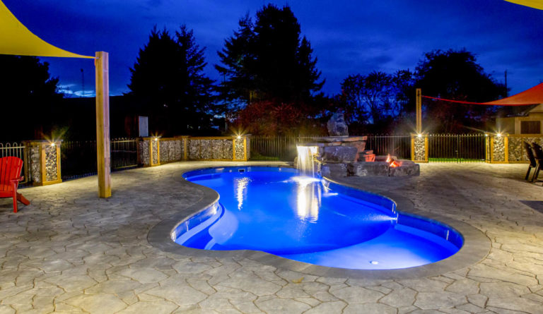 The Eclipse inground fiberglass pool illuminated at night with landscape lighting in Sapphire Blue